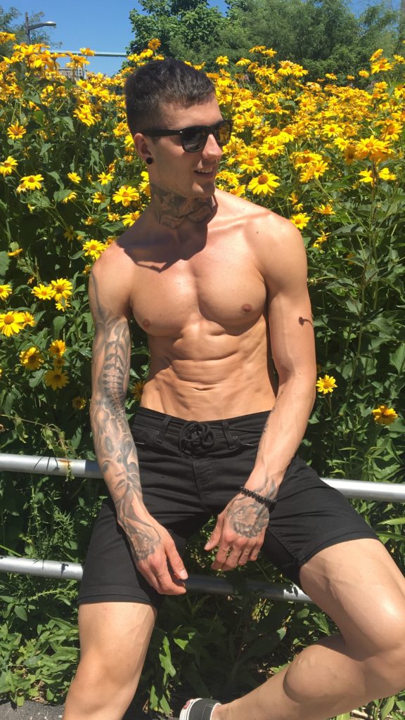 Jakob showing his lean stomach outside next to a bunch of yellow flowers.