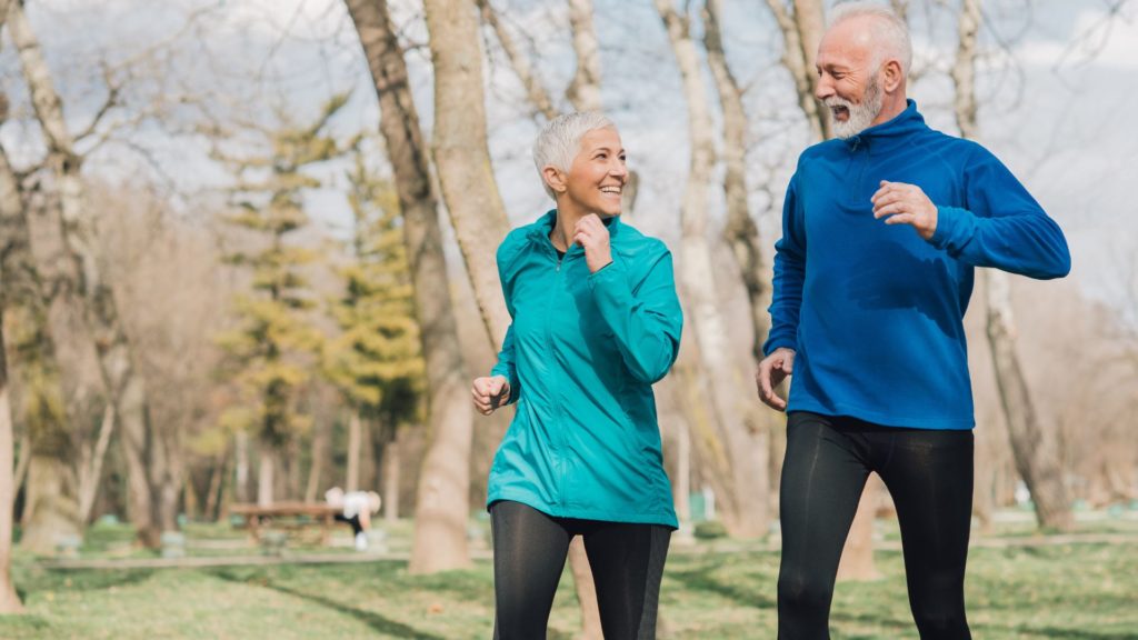 A middle-aged couple jogging in the park, smiling at eachother.