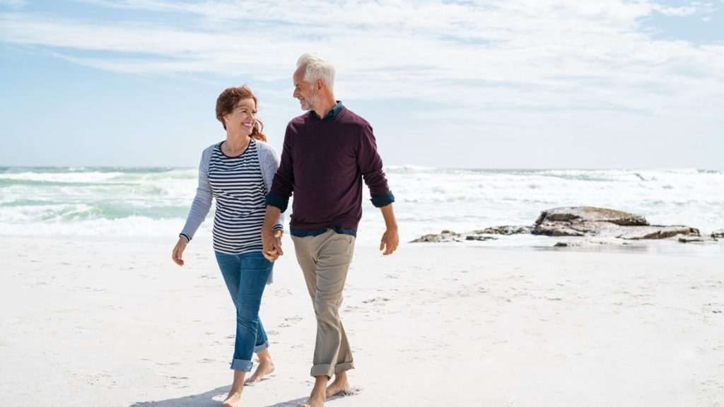 A middle-aged couple walking on the beach smiling at each other.