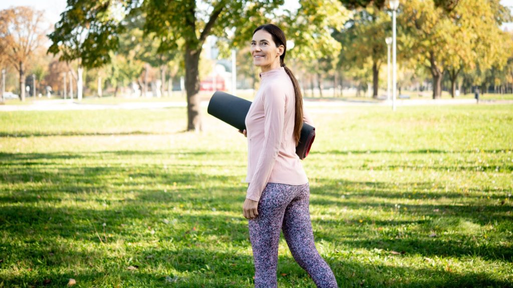 A middle-aged woman carrying a yoga mat through a park.